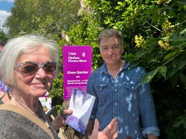 The image shows a man and woman standing in front of a sign for the Chelsea Flower Show, with the woman holding a pamphlet and the man wearing a denim shirt.
