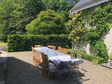 The image depicts a long table set for an outdoor meal on a sunny day, surrounded by lush greenery and a charming garden setting.