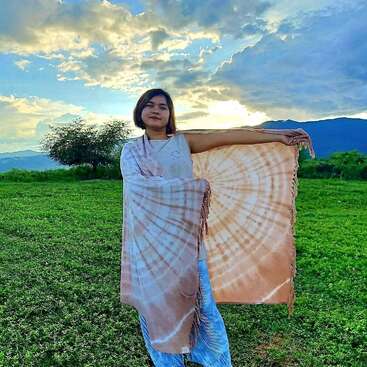 A woman stands in a green field during sunset, holding an orange tie-dye shawl. The sky is partly cloudy with dramatic sunlight and distant hills.