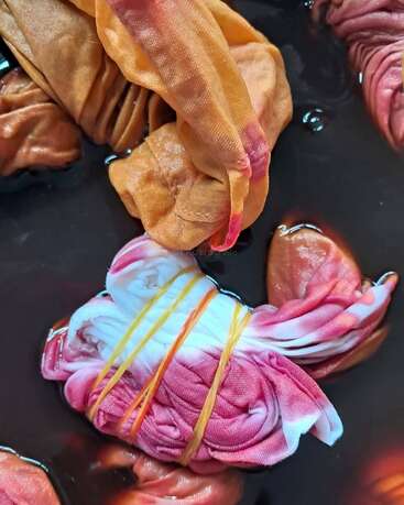Dieses Bild zeigt Stoffstücke, die verdreht und mit Gummibändern zusammengebunden sind und in dunkler Farbe einweichen – so entstehen bunte Batikmuster.
