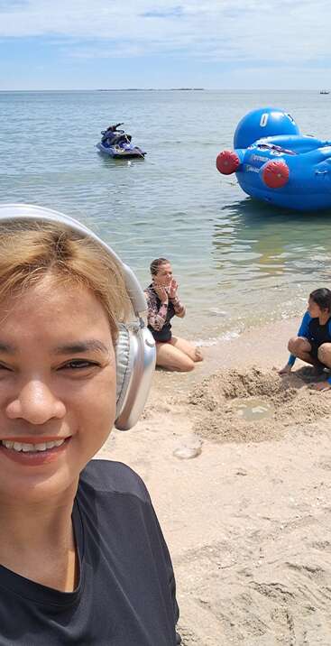 Drei Leute genießen einen sonnigen Tag am Strand. Einer lächelt mit Kopfhörern für ein Selfie, während die anderen am Wasser in der Nähe eines Jetskis spielen und sich treiben lassen.
