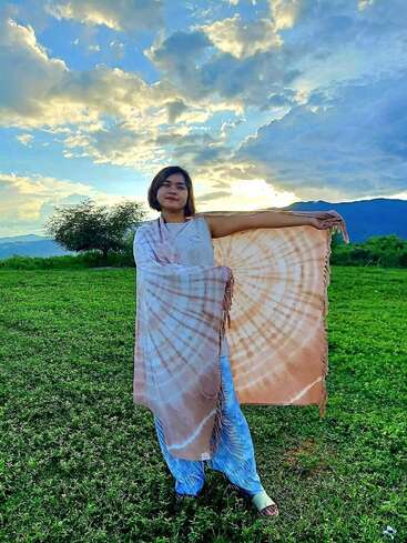 Uma mulher está em um campo verde ao pôr do sol, segurando um xale laranja com estampa tie-dye. O céu está parcialmente nublado, com uma luz do sol espetacular e colinas ao longe.