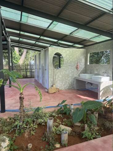 Dieses Bild zeigt eine Freiluft-Toilettenanlage mit einem Metalldach, Betonwaschbecken und mehreren Kabinen. Sie ist von Grünflächen und einem überdachten Garten umgeben.