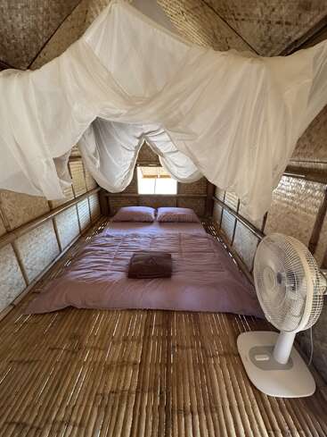 Ein gemütliches Schlafzimmer in einer Bambushütte mit einem rosafarbenen Bett, einem darüber drapierten Moskitonetz, einem kleinen Fenster, einem Ventilator, geflochtenen Wänden und einem rustikalen, natürlichen Ambiente.