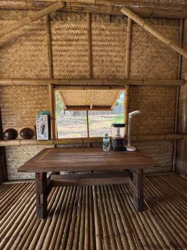 In einer rustikalen Bambushütte steht ein Holztisch mit einer Wasserflasche, einem Mixer und einer Lampe. Geflochtene Bambuswände, ein kleines Fenster und ein Bambusboden runden das Bild ab.
