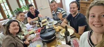 Seis personas están reunidas alrededor de una mesa disfrutando de una comida juntas y sonriendo a la cámara. La mesa está puesta con vino, pan, queso y embutidos.