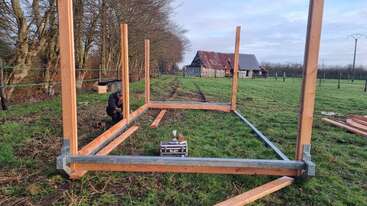 Una persona está construyendo una estructura de madera en un prado. Varias vigas de madera están colocadas en posición vertical, con herramientas y materiales esparcidos por el suelo. Al fondo se divisan graneros desgastados por el paso del tiempo.