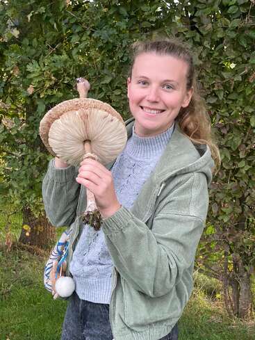 Una mujer sonriente está de pie al aire libre, sosteniendo una seta silvestre gigante con las láminas visibles, rodeada de follaje verde, y vestida con una chaqueta verde y un jersey gris.