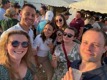 Un grupo de amigos disfruta de un evento al aire libre bajo el sol, sonriendo para una selfie con bebidas en la mano, rodeados de mucha gente bajo unas carpas blancas, en un ambiente festivo.