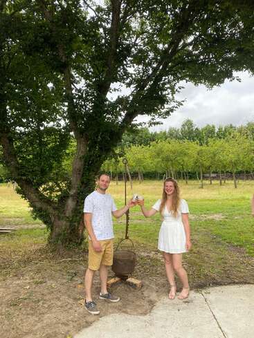Un hombre y una mujer brindan bajo un gran árbol en un frondoso parque. Sonríen, vestidos con ropa de verano, junto a una vieja olla de metal que cuelga de un gancho.