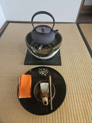This image depicts a traditional Japanese tea ceremony setup on tatami matting, including a cast iron teapot, bamboo whisk, tea bowl, cloth, and utensils.