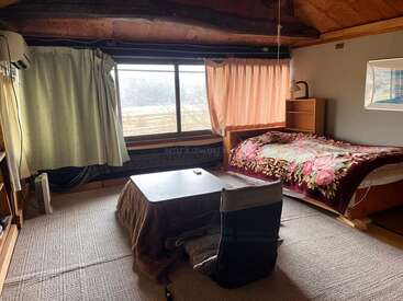 A cozy room with wooden ceiling beams, large windows, simple curtains, a single bed with a floral blanket, low table, chair, shelf, and soft lighting.