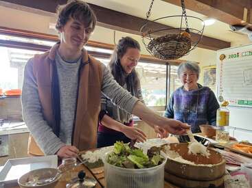 Three people are joyfully preparing food together in a bright kitchen, spreading rice while smiling, surrounded by fresh ingredients and a friendly, warm atmosphere.