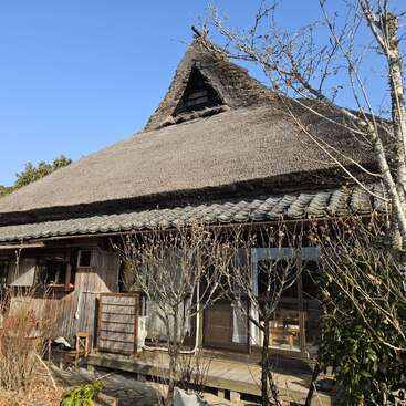A traditional Japanese house with a thatched roof and wooden exterior sits under a clear blue sky, surrounded by bare trees and natural greenery in the garden.