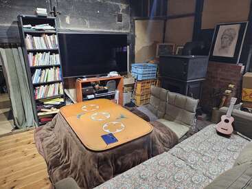 A cozy, cluttered living room with a kotatsu table, bookshelf, large TV, cushioned chairs, wood stove, crates, artwork, a ukulele, and patterned couch.