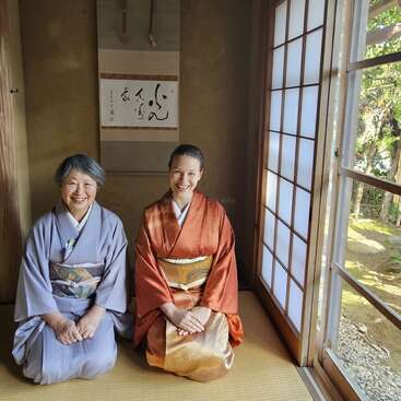 Two women in traditional kimonos sit on tatami mats in a Japanese tea room, smiling. Soft light enters through shoji screens, creating a peaceful atmosphere.