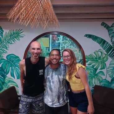 Trois amis souriants posent ensemble à l'intérieur. Derrière eux, on aperçoit une fresque murale représentant un feuillage verdoyant et une fenêtre ronde. L'ambiance est joyeuse et détendue, avec un décor tropical aux couleurs vives et un éclairage chaleureux.
