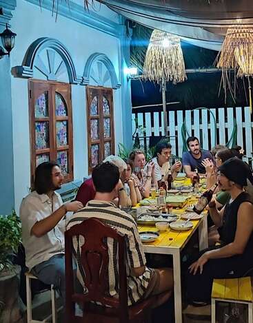 Un groupe d'amis dîne ensemble dans un restaurant en plein air, à la fois chaleureux et bien éclairé. Le décor se compose de murs bleus, de fenêtres en bois et de suspensions décoratives.