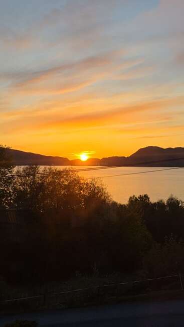 Un coucher de soleil à couper le souffle pare le ciel de teintes chaudes orange et jaunes, au-dessus d'un lac paisible, d'arbres se découpant en silhouette et de collines lointaines, créant ainsi une atmosphère sereine.