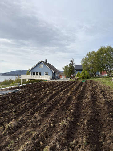 Une charmante maison bleue se dresse au bord d'un lac, sous un ciel nuageux. Au premier plan, la terre fraîchement labourée, prête à être ensemencée, est entourée d'arbres et de nature.