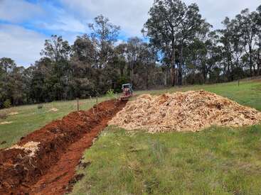 A freshly dug trench stretches through green grass near a forest. A small excavator is visible in the background, beside a large pile of mulch or wood chips.