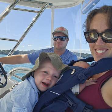 Una alegre familia disfruta de un soleado paseo en barco. La madre sostiene a un bebé en un portabebés, mientras un sonriente niño y su padre se relajan juntos en el agua.