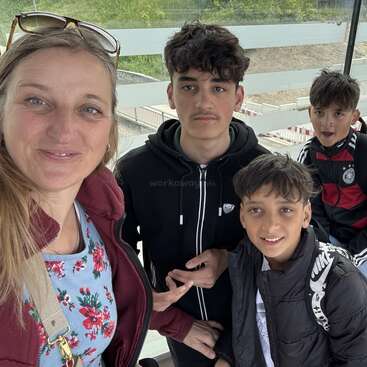 A woman and three boys smile for a selfie. They are outdoors, dressed in casual jackets, with greenery and construction barriers visible in the background.