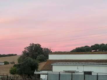 Eine ruhige ländliche Szene bei Sonnenuntergang mit einem weißen Haus mit Ziegeldach, üppigen Bäumen, sanften Hügeln und einem wunderschönen rosa und lila Himmel.