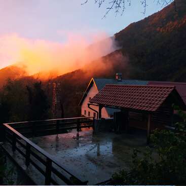 The image depicts a serene scene of a house with a tiled roof and a balcony, set against a backdrop of mountains and trees at sunset.