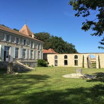 The image depicts a large stone house with a brown roof, set amidst a lush green lawn and surrounded by trees, with a clear blue sky above.