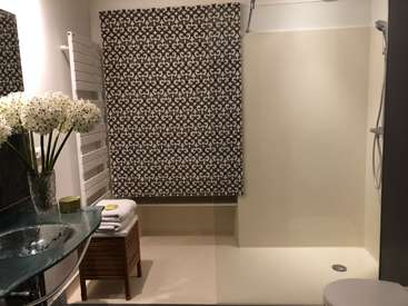 A imagem mostra um banheiro moderno com uma pia de vidro, toalhas brancas e uma área de chuveiro, com uma cortina estampada e um vaso sanitário branco no canto.