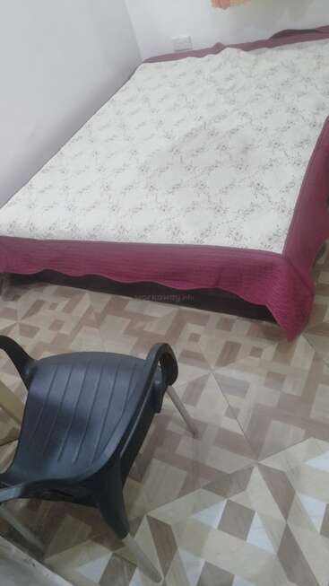 The image depicts a bedroom with a bed and chair, featuring a white and pink patterned bedspread, a black chair, and a geometric-patterned floor.