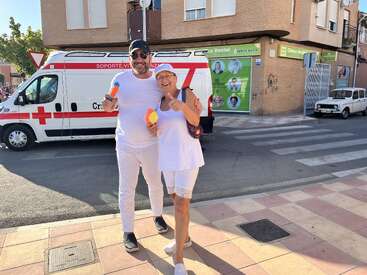 L'image représente un homme et une femme debout sur un trottoir, posant pour une photo, avec une ambulance à l'arrière-plan, sur fond de bâtiment en briques.