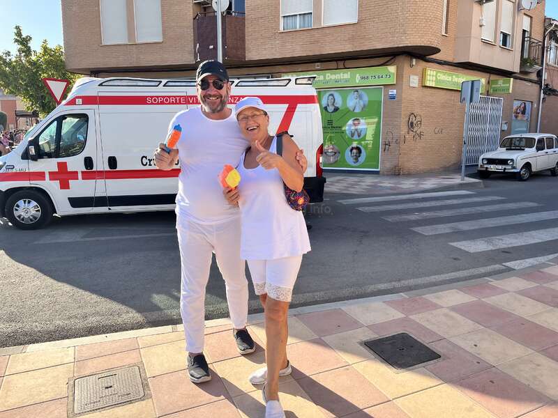 Das Bild zeigt einen Mann und eine Frau, die auf einem Bürgersteig stehen und für ein Foto posieren, mit einem Krankenwagen im Hintergrund und einem Backsteingebäude als Kulisse.