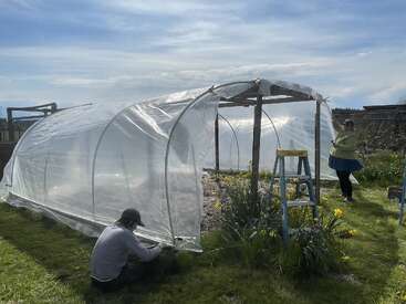 Das Bild zeigt zwei Personen, die ein Gewächshaus bauen. Die eine kniet, um eine Plastikfolie an der Konstruktion zu befestigen, die andere steht daneben und hält eine weitere Folie.