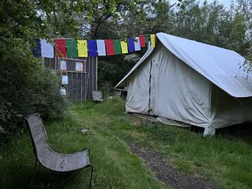 Das Bild zeigt eine ruhige Umgebung im Freien mit einem weißen Zelt, bunten Gebetsfahnen und einem Korbstuhl, umgeben von üppigem Grün und einer Holzkonstruktion.