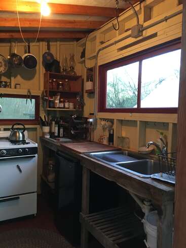 Das Bild zeigt eine gemütliche Küche mit einem weißen Herd, einer hölzernen Arbeitsplatte und einer Spüle, mit verschiedenen Kochutensilien und einem Fenster, das den Blick auf die Bäume draußen freigibt.