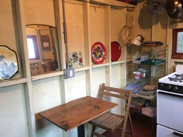 Das Bild zeigt eine kleine Küche mit einem Holztisch und -stuhl, einem weißen Herd und verschiedenen Kochutensilien und Geschirr an den Wänden und auf den Regalen.