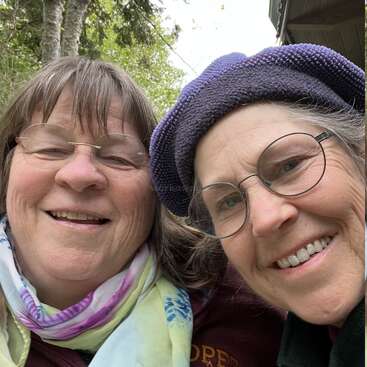 Das Bild zeigt zwei Frauen, die in die Kamera lächeln, mit Bäumen und einem Gebäude im Hintergrund, was auf eine Tageszeit im Freien hindeutet.