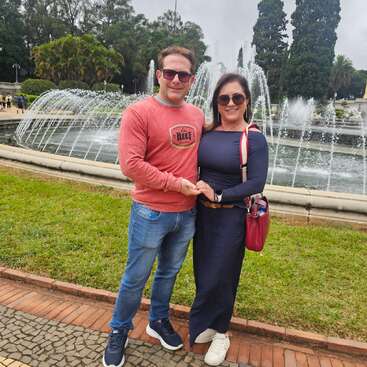 Um casal sorridente está de mãos dadas em frente a uma grande fonte, cercado por vegetação e árvores altas; os dois usam óculos escuros e roupas casuais.