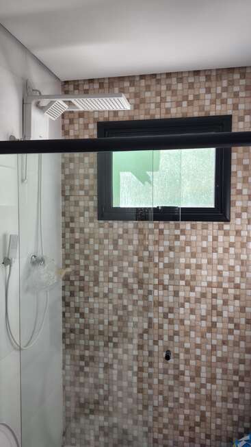 Esta imagem mostra uma área de banho moderna com um chuveiro tipo chuva quadrado, divisória de vidro, azulejos de mosaico bege nas paredes e uma janela fosca com moldura preta que deixa entrar luz natural.