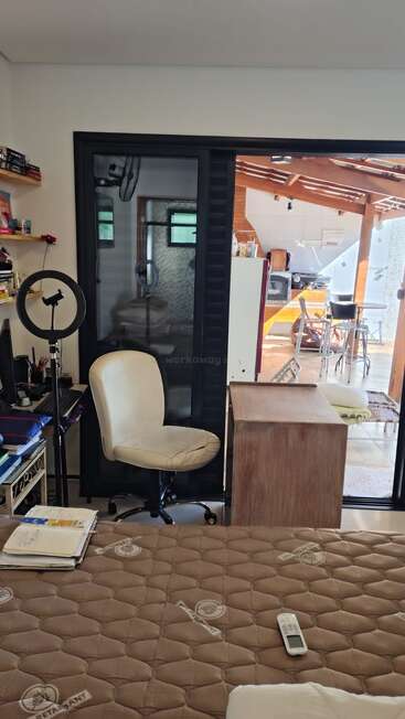 Um quarto aconchegante com uma mesa, cadeira de escritório, luz anelar, computador, colchão com livros e controle remoto. As portas de vidro deslizantes dão acesso a uma cozinha aberta e bem iluminada.