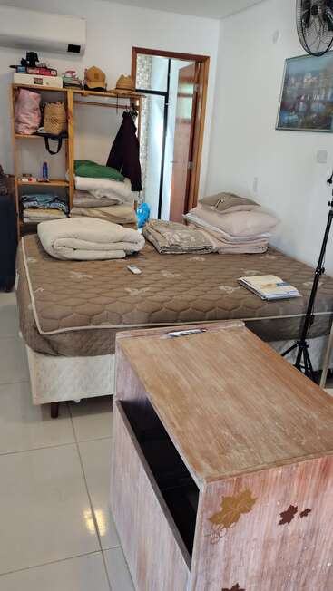 Um quarto aconchegante com uma cama, colchão marrom, roupa de cama dobrada, prateleiras abertas, roupas, uma mesa de madeira, ventilador de parede, porta aberta e vários objetos pessoais.