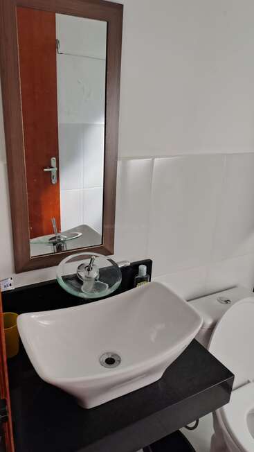 Esta imagem mostra um banheiro com um espelho retangular, uma pia de apoio moderna, uma torneira de vidro, um copo amarelo, um dispensador de sabonete e um vaso sanitário branco ao lado.