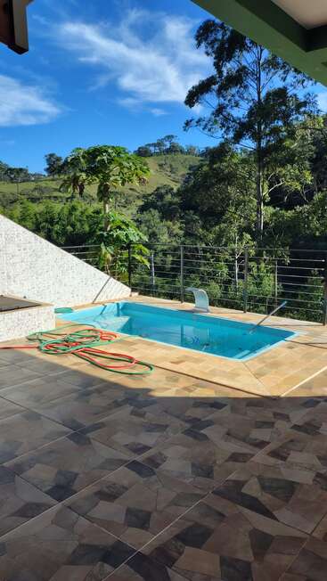Uma piscina ao ar livre tranquila fica em um pátio de azulejos, cercada por uma vegetação exuberante e colinas. Uma mangueira de jardim está enrolada ali perto, sob um céu azul brilhante.