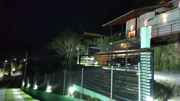 Uma casa moderna no alto de uma colina fica iluminada à noite. Um portão de metal, uma cerca e um caminho com luzes brilhantes levam até a entrada principal. Um ambiente tranquilo.