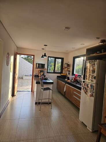 Esta imagem mostra uma cozinha moderna com armários de madeira, uma geladeira coberta de fotos, uma ilha com uma cadeira e a luz do sol entrando pelas janelas e portas abertas.