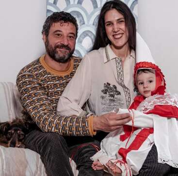 L'image représente une famille de trois personnes, composée d'un homme, d'une femme et d'un bébé, assis avec un chien sur un canapé, contre un mur blanc à motifs bleus.