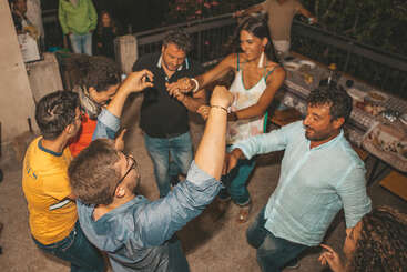 A group of people are joyfully dancing together in a circle outdoors at night, surrounded by tables with food, enjoying a lively and festive gathering.