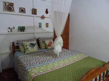 The image depicts a cozy bedroom with a wooden bed frame, a vibrant quilted comforter, and a white canopy, set against a clean white wall with decorative shelves.